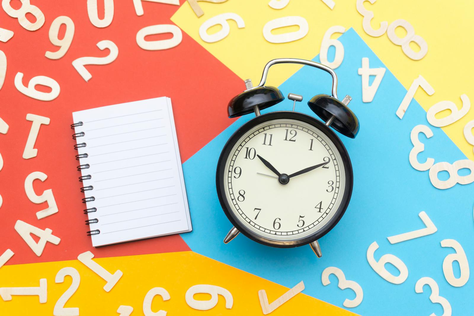 A flat lay of a classic alarm clock with a blank notebook on a colorful background surrounded by numbers.