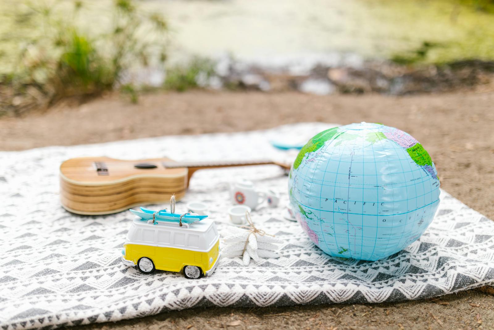 A cozy outdoor picnic setup featuring a globe, toy van, and guitar on a blanket by the water.